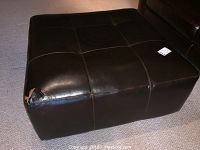 Large square dark brown leather ottoman with tufted stitching, slight sheen, and a circular indentation in center, placed on carpet.