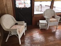 Two white wicker chairs and two small wicker tables in a sunroom with wooden floors and paneled walls.