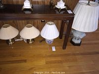 Wide view of six assorted lamps of various sizes and designs on hardwood floor next to wooden furniture.