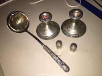 Two sterling silver weighted candlesticks with decorative engravings, small salt and pepper shakers, and a ladle with ornate floral handle on a flat light surface.