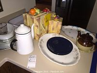 A collection of serving items including white enamel teapot, brown ceramic teapot, floral platter, blue plate, and three wooden decorative storage boxes with fruit.