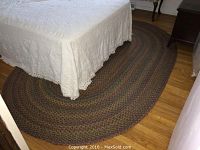Oval braided rug shown mostly under a bed, displaying its multicolored pattern and oval shape on hardwood floor.