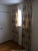 Curtains with floral fabric hung on either side of a tall narrow window with a white window blind inside the frame.