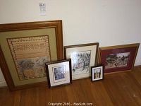 Five framed artworks arranged on the floor against a white wall showing variety of sizes and styles including sampler and painted scenes.