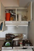 Photo showing kitchen cabinet open with large orange pitcher, hand coffee grinder, glass pitchers and jars and white insulated thermos inside. Below cabinet counter has wooden spice racks, plastic water filter pitcher, kitchen timer, spice jars and other kitchen items arranged.