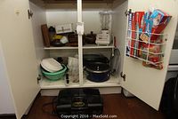 Kitchen cabinet with mix of pots, pans, bowls, blender, glass casserole, cutting boards, and storage rack holding multiple kitchen wraps on door.