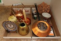 Overview photo showing lot contents including copper tea kettle, candle holder with candle, brass watering can, decorative copper wall hanging, enamel decorative plate on stand, copper pan with black handle, and several small pottery pieces.