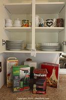 Cupboard contents showing mugs, cream and sugar sets, mixed plates, dipping set, and boxed kitchen tools on countertop below