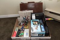 Photo showing the full lot including brown leather briefcase or portfolio, Samsung digital photo frame box, Bushnell binoculars case, various office supplies in a box on carpeted floor.