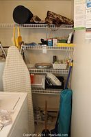 Photo of wire shelving with household items including ironing board, handheld vacuum, cleaning supplies, and utility bags on top shelf.