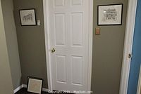 Two framed ink prints of street scenes hanging and leaning against wall in hallway corner.