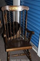 Front view of dark stained maple wood rocking chair with spindle back and armrests, showing floral design on upper backrest.