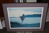 Framed artwork showing a girl walking on train tracks by water with scenic background and hills.