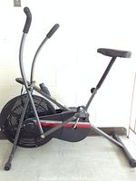 Side angle showing entire black and gray Wind Master exercise bike with dual moving handles, large flywheel, padded adjustable seat, and foot pedals with straps.