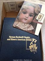 Blue hardcover book titled 'Norman Rockwell Classics and Historic American Stamps' with gold embossed title and illustration lying on a carpet surface alongside a booklet featuring a vintage doll.