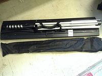 Accolade Duet projection screen and stand folded into black metal case with carrying bag alongside on the floor.