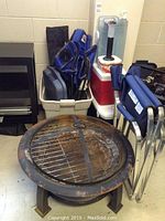 Metal fire pit with rusted surface, removable grill, and mesh spark screen on the ground with cooler and chairs behind