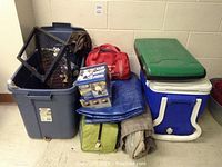 Photo of all camping gear in a group including cooler, tarps, tent bag, camp chair, first aid kit, lamp box, and metal frame.