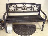 Front view of black metal garden bench with decorative backrest and rubber backed door mat beneath, with bocci game case visible on the left front side.