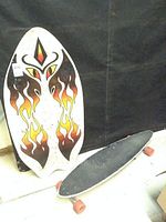 Photo showing the beach skim board with flame design leaning against the black backdrop next to the longboard skateboard with a black grip tape top and orange wheels laying on the floor.