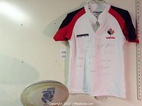 Photo of signed Canada Rugby 7's white jersey with red and black sleeves hanging on wall, showing multiple illegible player signatures.