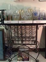 Metal wine rack with black granite top, featuring intricate scroll metalwork and capacity for 20 bottles, photographed with assorted glasses and accessories on and around it.