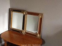 Photo of two decorative wood trim wall mirrors resting against a wall on a wooden table surface. Mirrors have gold-toned carved frames with corner accents.
