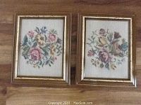 Two framed needlepoint floral artworks displayed side by side on a wooden surface.