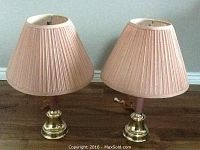 Pair of dusty rose table lamps with pleated fabric shades and brass-tone bases sitting on wooden floor against white wall.