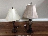 Photo showing two table lamps on a wooden surface against a plain wall, one shorter and one taller with distinct fabric lampshades and metal bases.