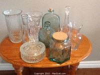 Nine assorted glass vases, bottles, and containers of various shapes and sizes on a wooden table.