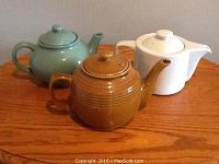 Photo showing a collection of three ceramic teapots in different colors (green, brown, white) placed on a wooden surface.