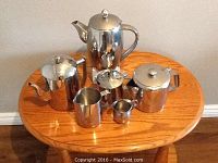 Six stainless steel coffee service pieces arranged on a small wooden table, including coffee pots, creamer, sugar bowl, and small pitcher.