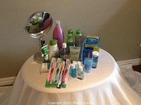 Photo showing various unopened health and beauty supplies on a round table including toothbrushes, toothpaste, shampoo, lotion, hand sanitizer, lip balm, deodorant, and a makeup mirror.