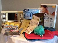 Shows king size heating pad box, makeup mirror with lights, bath salts, perfumed soaps, nail accessories, green bath pouf, and a red hot water bottle on a surface.