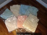 Seven individually packaged knitted baby clothing sets in various pastel colors arranged on a wooden floor.