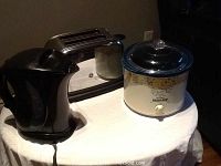 Photo showing three kitchen appliances on a small round table: black Proctor Silex electric kettle on the left, stainless steel GE toaster in the center, and Rival small crock pot with floral design and dark lid on the right.