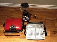 George Foreman red electric grill with metal handle next to an Oster blender with clear 1.25L pitcher and 12-speed dial, placed on floor.