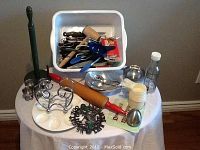 Full view of assorted kitchen tools on small round table including egg cup holder, timer, rolling pin, napkin holder, serving bowl with tongs, measuring cups, glass bottle, and utensil bin.
