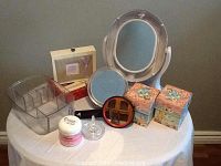 Photo of vanity set including Revlon electric mirror with bulbs, acrylic makeup organizer, two decorative floral boxes, a small red-framed mirror, stationary sets, jar of Ocean Sleep cream, and a jewel cleaner on a round table.