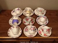 Photo shows nine assorted bone china tea cups on their matching saucers arranged in rows on a wooden surface. Each cup has distinct floral patterns with various colors including pink, purple, red, and yellow.