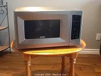 Photo shows Kenmore stainless steel microwave oven placed on a round wooden table against a wall, viewed from the front and slightly to the left side.