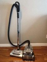 Photo of the VC336EB1 vacuum cleaner showing the main unit with clear removable canister, black hose, adjustable metal wand, and floor head on hardwood floor against beige wall.