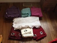 Photo of folded linens sets in purple, green, and pink hues arranged behind a lacey white tablecloth and grommeted drapes in burgundy and cream colors on a wooden floor.