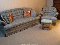 Photo shows matching Chesterfield style sofa and lounge chair, both upholstered in tufted blue-gray fabric with rolled arms. Several decorative pillows on sofa. Non-matching blue fabric sliding stool with wooden base in front of chair.
