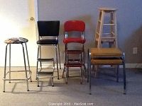 Photo showing six stools: two retro fold down stepping stools (black and red padded seats), two wooden kitchen stools, one upholstered stool with patterned seat, and two stacked general purpose stools.