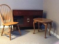 Photo shows a dark wood cabinet with one wide drawer and two sliding glass doors, a light wood side table with curved legs, and a light wood spindle back chair on a carpeted floor.