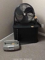 Photo showing a black TV stand with a pop-open door and two interior shelves, a large black Honeywell floor fan placed on top, and a silver radio cassette player on the carpet in front.