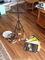 Photo showing the black metal adjustable music stand, two tambourines, two small wooden percussion instruments, and pile of assorted sheet music including books and loose sheets.