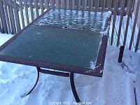 Angled view of square wrought iron patio table with green-finish glass top, umbrella hole, snow-dusted